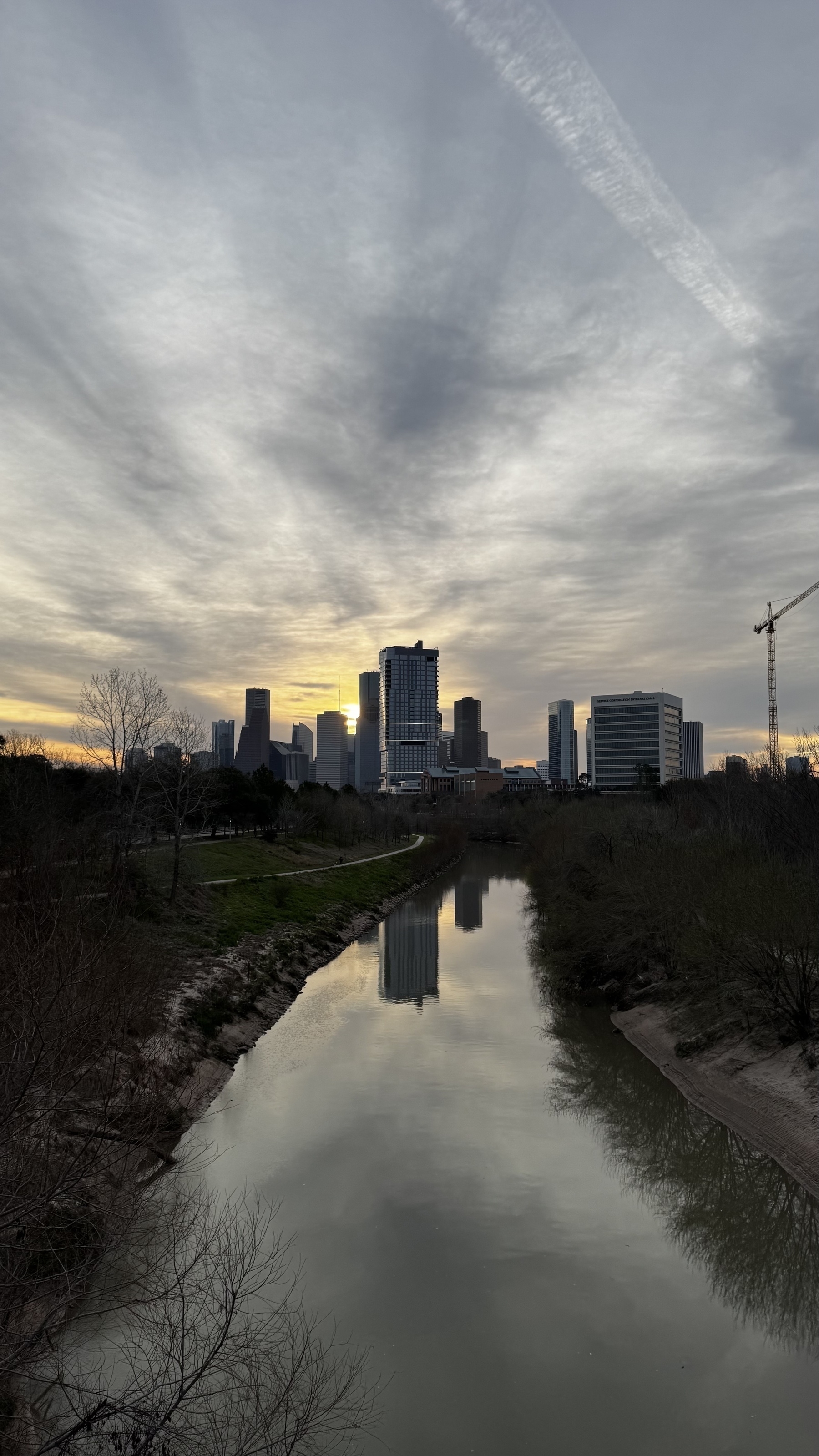 A cool partly cloudy morning, Houston skyline with the sun just trying to poke over the buildings. A calm still buffalo bayou in the foreground. 